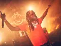Rapper mit Dreadlocks und orangefarbenem T-Shirt singt mit erhobenen Armen und Mikrofon. Die Bühne ist in warmes Licht getaucht, perfekt für energiegeladene Hip-Hop-Songs.