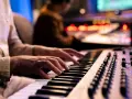 Hände spielen Synthesizer-Tasten, während im Hintergrund ein Tontechniker an einem Mischpult arbeitet. Die Szene fängt die Entstehung eines neuen ep album ein.