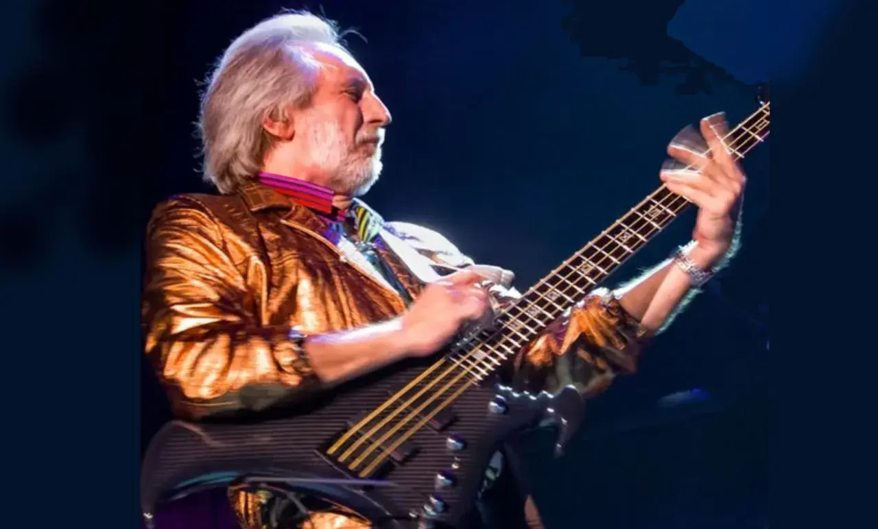 Ein Mann mit grauem Haar und Bart spielt Bassgitarre. Er trägt eine glänzende goldene Jacke und bunte Krawatte. Er ist der beste Bassist der Welt.