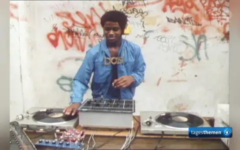 DJ Kool Herc, der oft als derjenige gilt, der Hip Hop erfunden hat, legt auf. Graffiti zieren die Wand im Hintergrund.