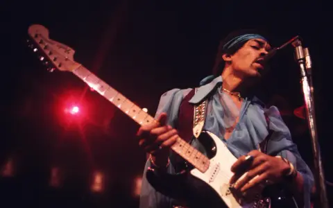 Jimi Hendrix, einer der besten Gitarristen, spielt auf der Bühne seine weiße Fender Stratocaster. Rotes Licht beleuchtet die Szene.