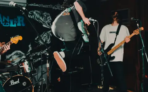Ein Gitarrist springt auf der Bühne, die Energie des Punk der 70er Jahre ist spürbar. Ein Bassist spielt im Hintergrund.
