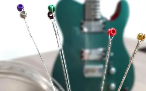 Vier Gitarrensaiten mit farbigen Enden (lila, grün, rot, gold) liegen vor einer grünen E-Gitarre. Die besten E-Gitarren-Saiten für deinen Sound.