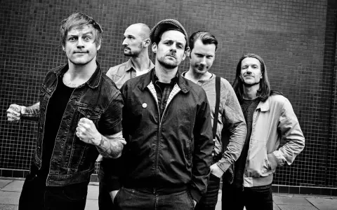 Fünf Männer stehen vor einer gemusterten Wand. Sie sehen aus wie eine energiegeladene Punkband, bereit, die Bühne zu rocken.
