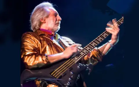 Ein Mann mit grauem Haar und Bart spielt Bassgitarre. Er trägt eine glänzende goldene Jacke und bunte Krawatte. Er ist der beste Bassist der Welt.