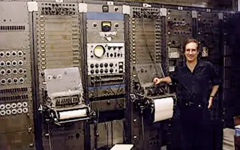 Mann steht neben einem riesigen, frühen Computer, dem ersten Synthesizer der Musikgeschichte.