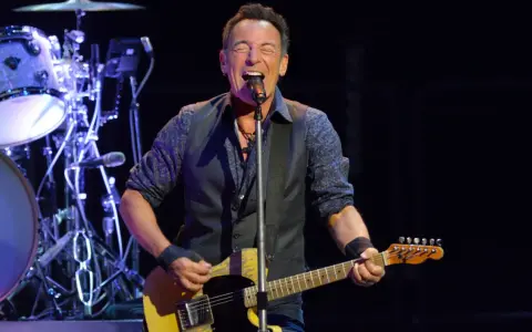 Bruce Springsteen singt und spielt seine ikonische Bruce Springsteen Gitarre auf der Bühne, während die Band im Hintergrund spielt.