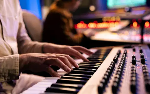 Hände spielen Synthesizer-Tasten, während im Hintergrund ein Tontechniker an einem Mischpult arbeitet. Die Szene fängt die Entstehung eines neuen ep album ein.