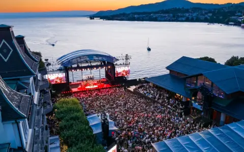 Ein großes Publikum feiert bei Sonnenuntergang ein Konzert am See. Die Bühne des Jazz Montreux Festivals ist beleuchtet, im Hintergrund der Genfersee.