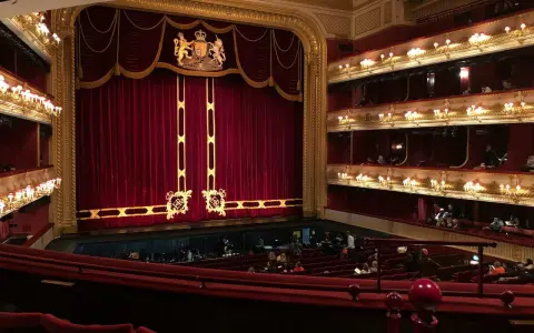 Ein prunkvolles Opernhaus mit roten Samtvorhängen und goldenen Verzierungen. Die Logen sind mit Lichtern geschmückt, und im Parkett sitzen Zuschauer.