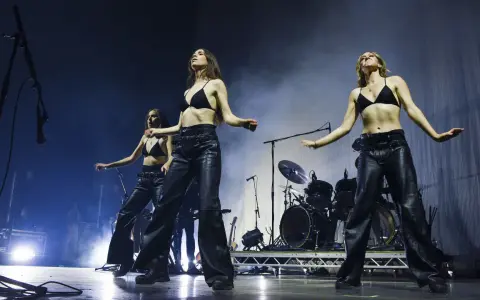 Drei Frauen in Lederhosen und BHs performen auf der Bühne, ein Schlagzeug im Hintergrund. Sie sind wie eine moderne Metal-Band.