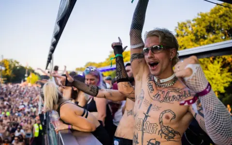 Feiernde Menschenmenge bei einem Festival, die zu **techno music** tanzt. Ein Mann mit Tattoos und Sonnenbrille jubelt.