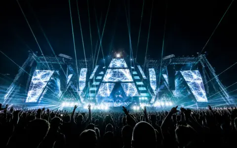 Eine riesige Menge jubelt bei einem EDM-Konzert. Die Bühne ist mit geometrischen Mustern und blauen Lichtern beleuchtet, was die Bedeutung von EDM-Musik für die Fans unterstreicht.