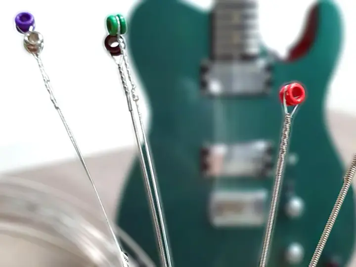 Vier Gitarrensaiten mit farbigen Enden (lila, grün, rot, gold) liegen vor einer grünen E-Gitarre. Die besten E-Gitarren-Saiten für deinen Sound.