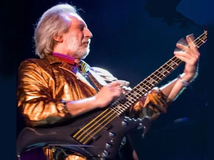 Ein Mann mit grauem Haar und Bart spielt Bassgitarre. Er trägt eine glänzende goldene Jacke und bunte Krawatte. Er ist der beste Bassist der Welt.