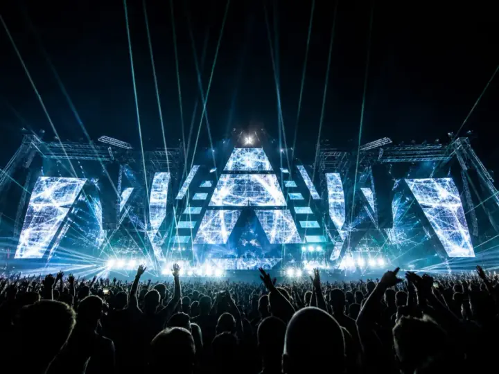 Eine riesige Menge jubelt bei einem EDM-Konzert. Die Bühne ist mit geometrischen Mustern und blauen Lichtern beleuchtet, was die Bedeutung von EDM-Musik für die Fans unterstreicht.